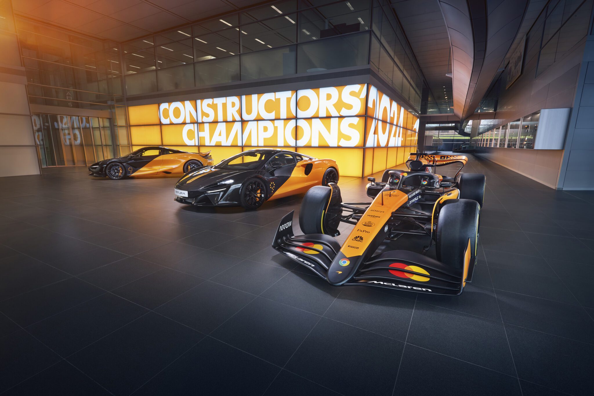 Three sleek orange and black McLaren performance cars, including the 750S and the 2024 MCL38, are parked in a modern garage. A sign in the background proudly reads "CONSTRUCTORS CHAMPIONS," highlighting their engineering excellence.