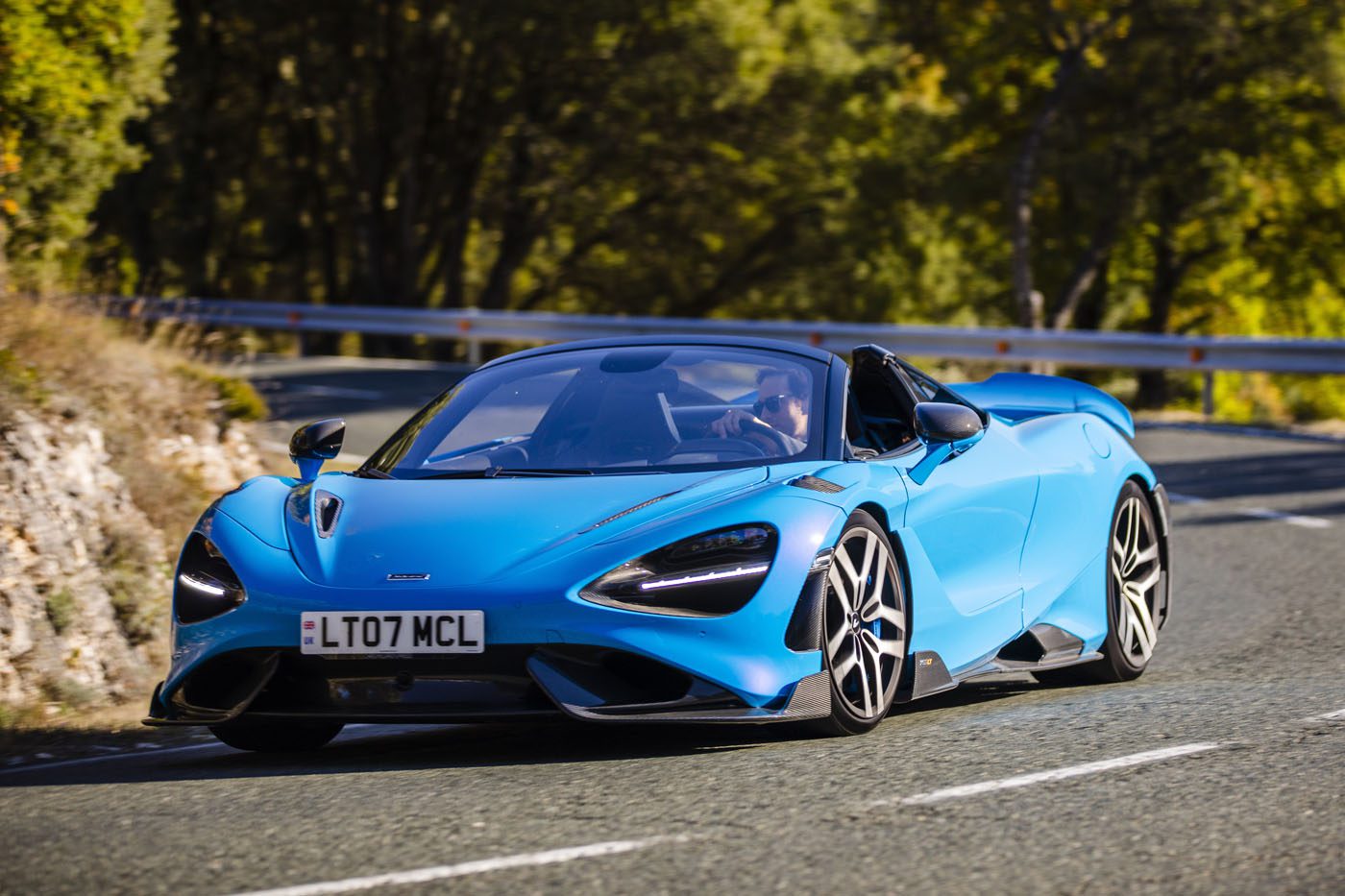A sleek blue McLaren navigates a curved road flanked by lush trees, embodying the allure of LT Cars.