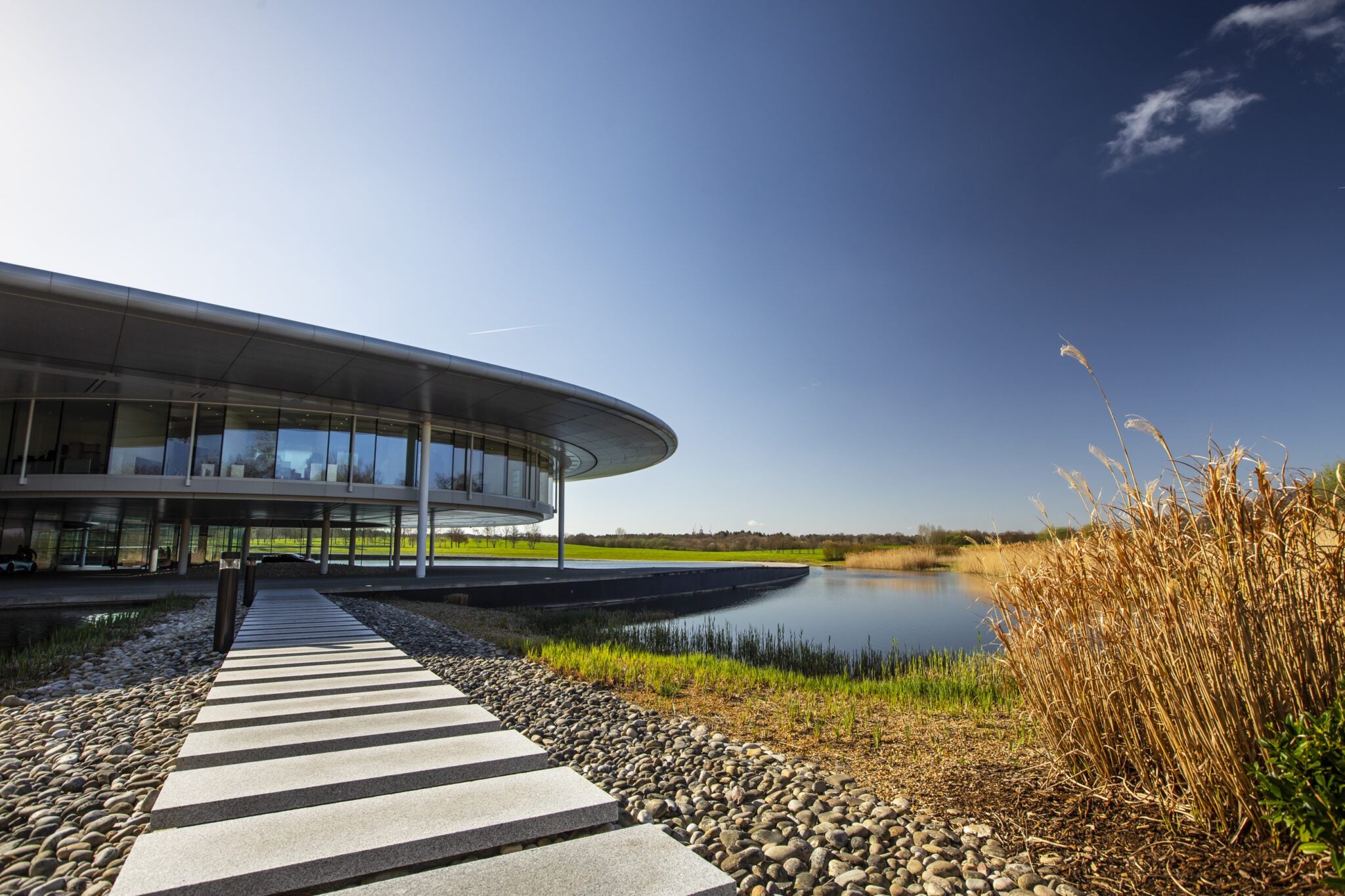 A sleek pathway leads to a modern, circular glass building by a serene pond under a clear blue sky with scattered clouds, symbolizing McLaren's brand reinvention—a fusion of innovation and elegance akin to their new future SUV.