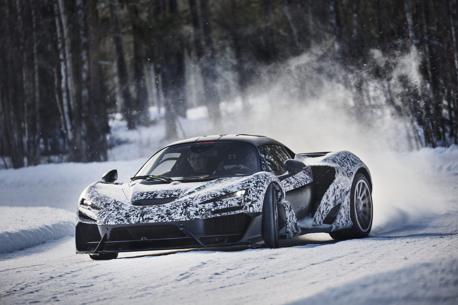 A McLaren W1 sports car with camouflage wrap speeds along a snow-covered ice track, kicking up snow as it rounds a curve during winter testing in a forested area.