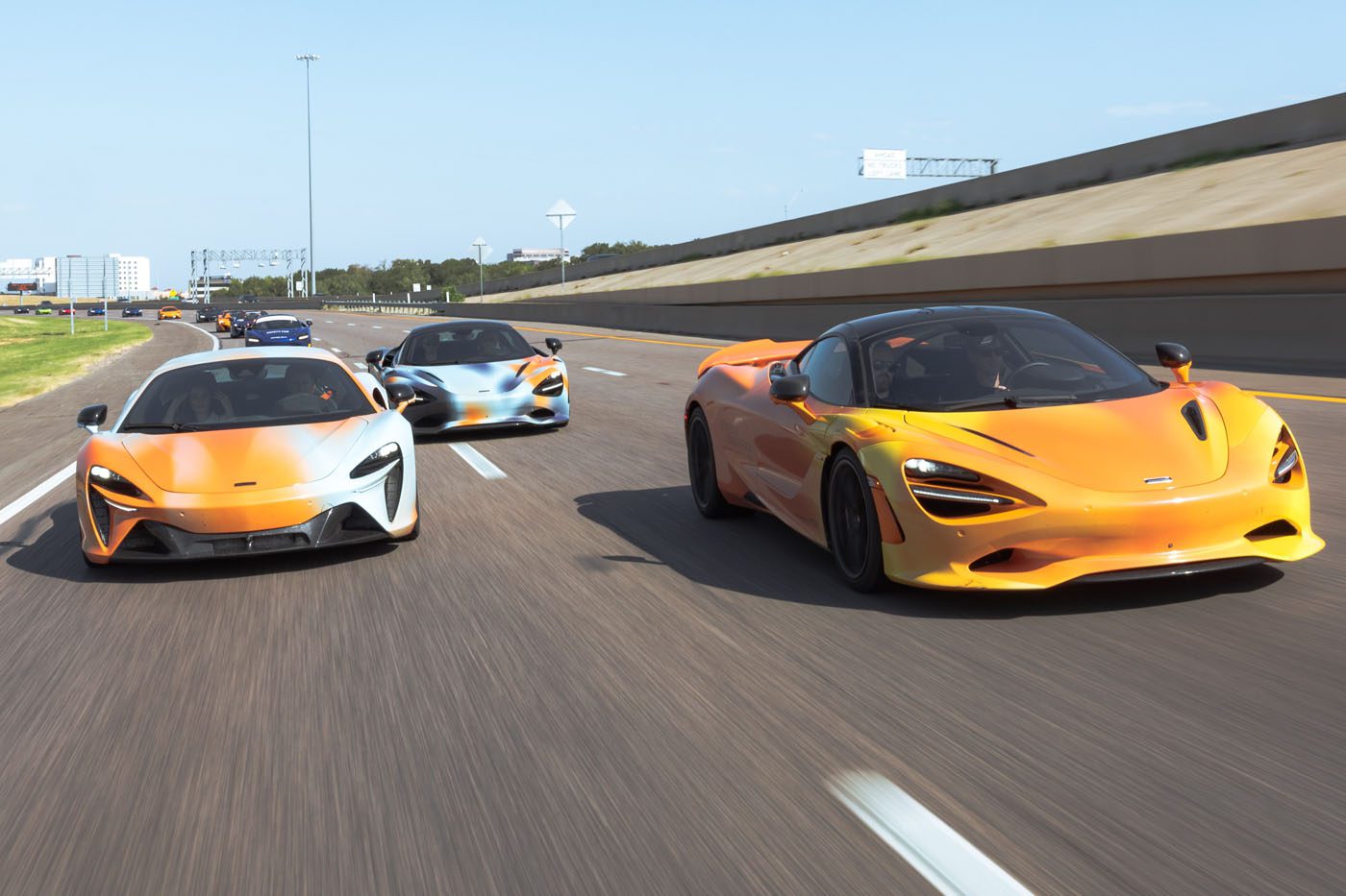 Three orange McLaren sports cars drive side by side on a multi-lane highway under a clear sky, embodying the spirit of a States of Endurance road trip.