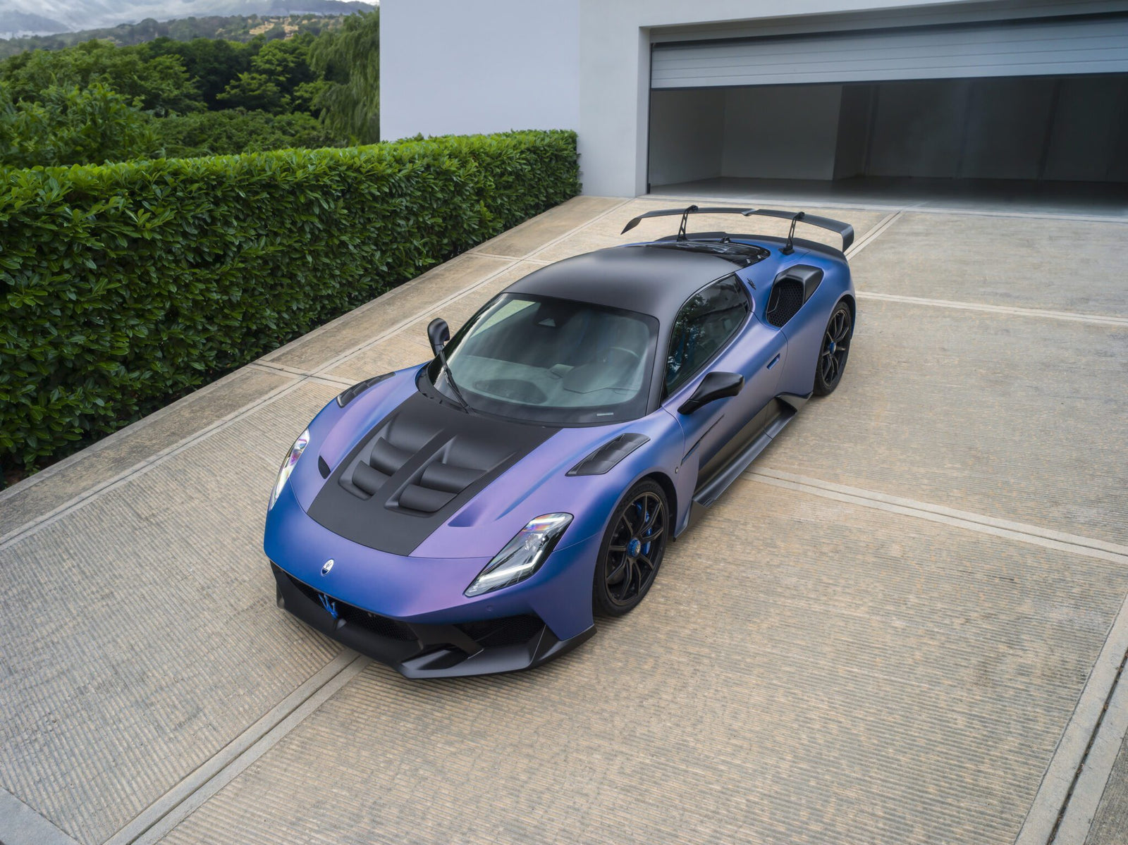 The sleek Maserati GT2 Stradale, with its striking blue finish and black roof, is parked on a driveway beside a white garage and lush green hedge.