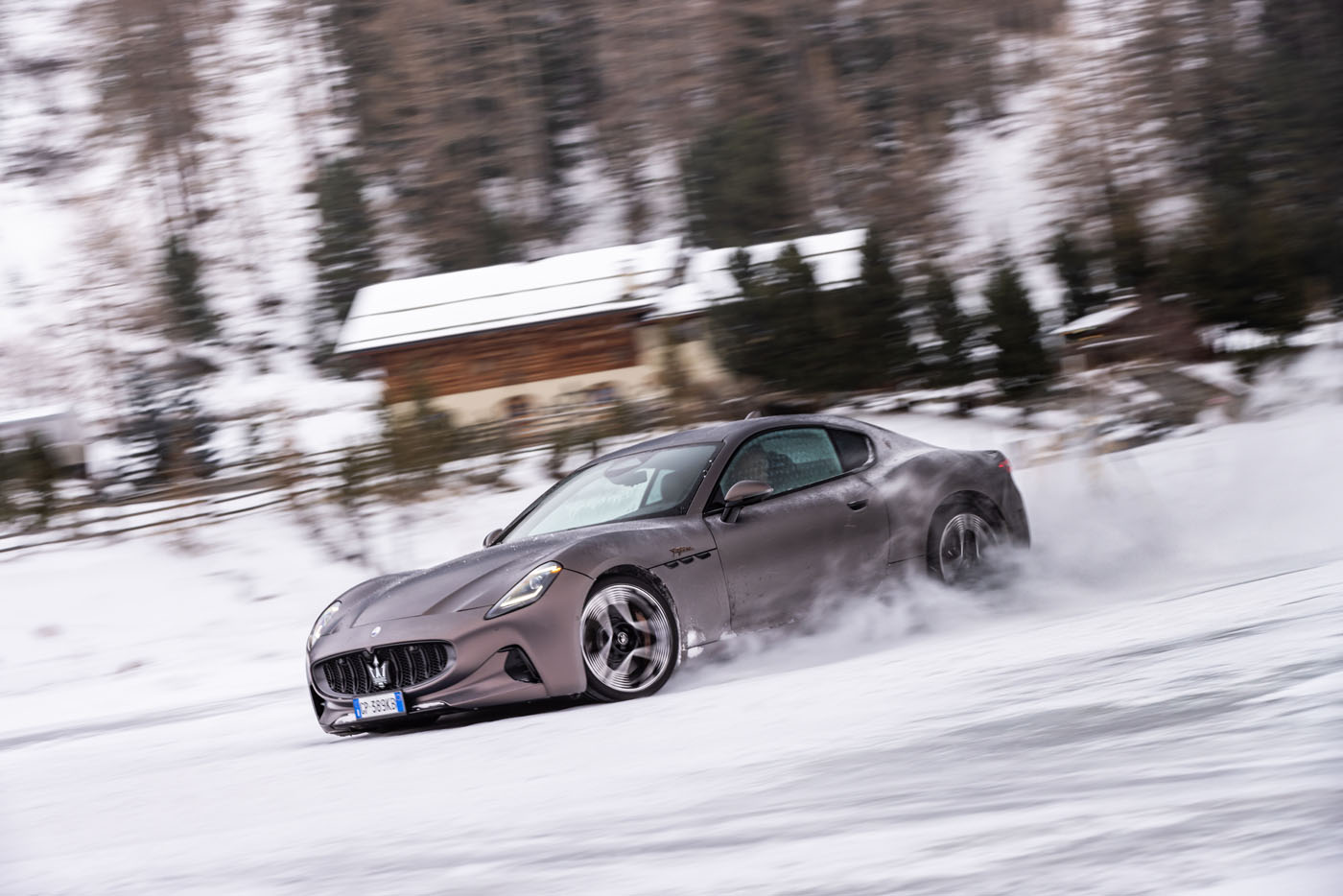 A gray Maserati GranTurismo elegantly drifts on a snowy road, creating a spray of snow. Trees and a wooden building are visible in the background.