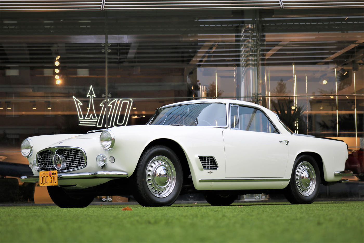 A vintage white Maserati 3500GT is elegantly parked on the green grass in front of a glass building adorned with a "110" logo, showcasing its timeless Classiche charm.