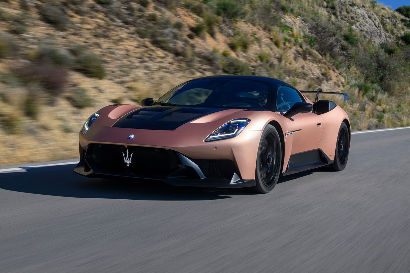 A bronze Maserati MC20 Cielo sports car drives on a paved road with hills and greenery in the background, showcasing its style at the Motor Valley Fest.