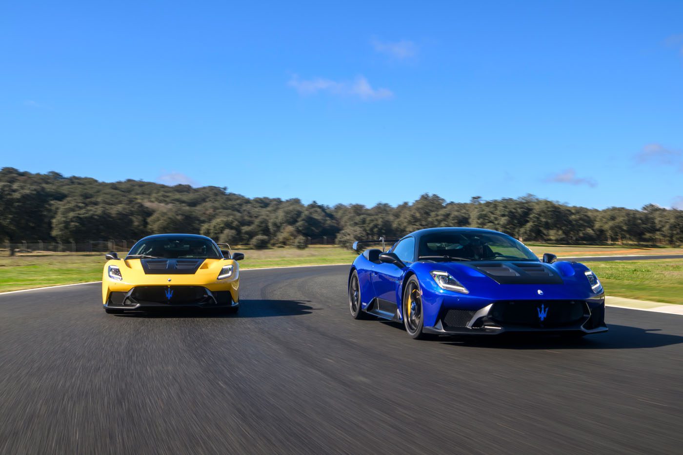 Two sports cars, a yellow Maserati and a blue GT2 Stradale, drive side by side on a paved road with lush greenery and the clear blue sky of Spain in the background.