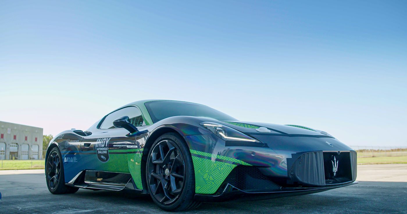 A sleek, dark-colored Maserati MC20 is parked outdoors on a clear day, its visible logos and graphics shining under the sun, hinting at its autonomous driving capabilities.