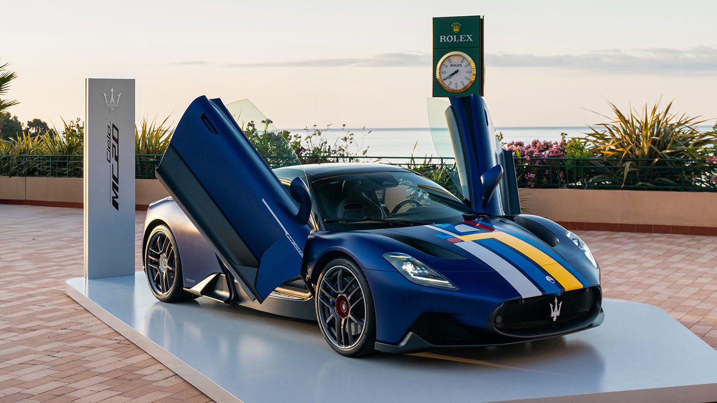 A blue Maserati MC20 Cielo with upward-opening doors is displayed on a terrace near the sea, evoking the elegance of the Rolex Monte-Carlo Masters. A distinguished Rolex clock stands nearby, adding to the scene's luxury.