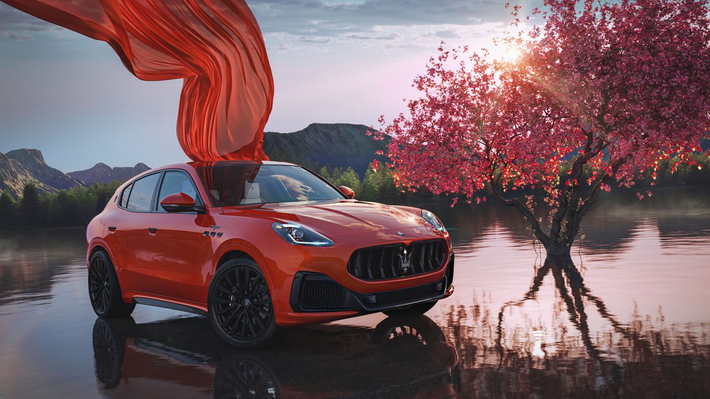 An orange 2025 Maserati Grecale SUV is parked beside a blooming pink tree near a reflective lake, with a red cloth draped over part of the car.