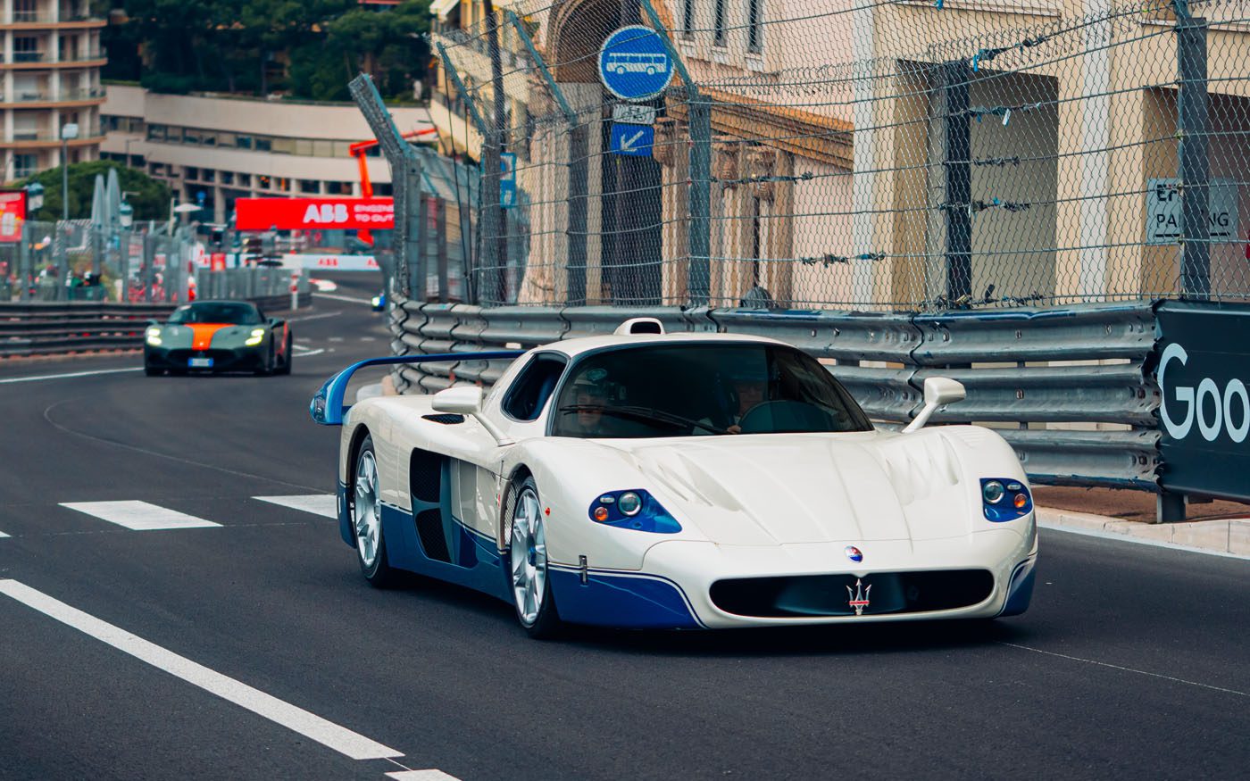 A white and blue Maserati MC12 supercar speeds along a fenced city racetrack during the Monaco E-Prix, with another sports car close behind.