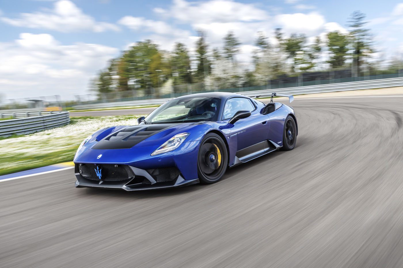 A blue Maserati GT2 Stradale speeds around a racetrack on a clear day, with trees and a blue sky in the background, capturing the thrill of the Maserati Driving Experience.