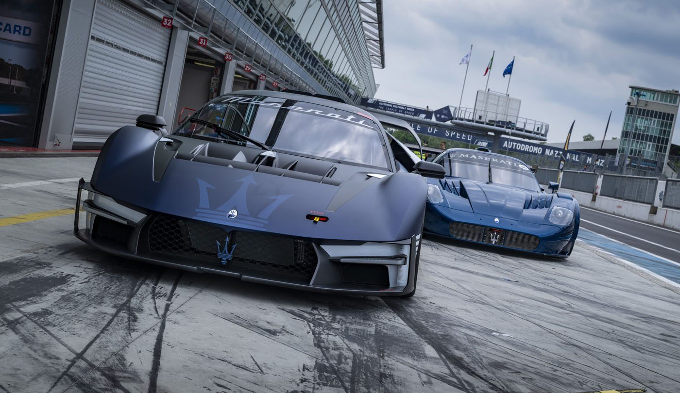 Two blue Maserati MCXtrema sports cars are parked on a Monza racetrack pit lane, with a garage and racetrack facilities visible in the background.