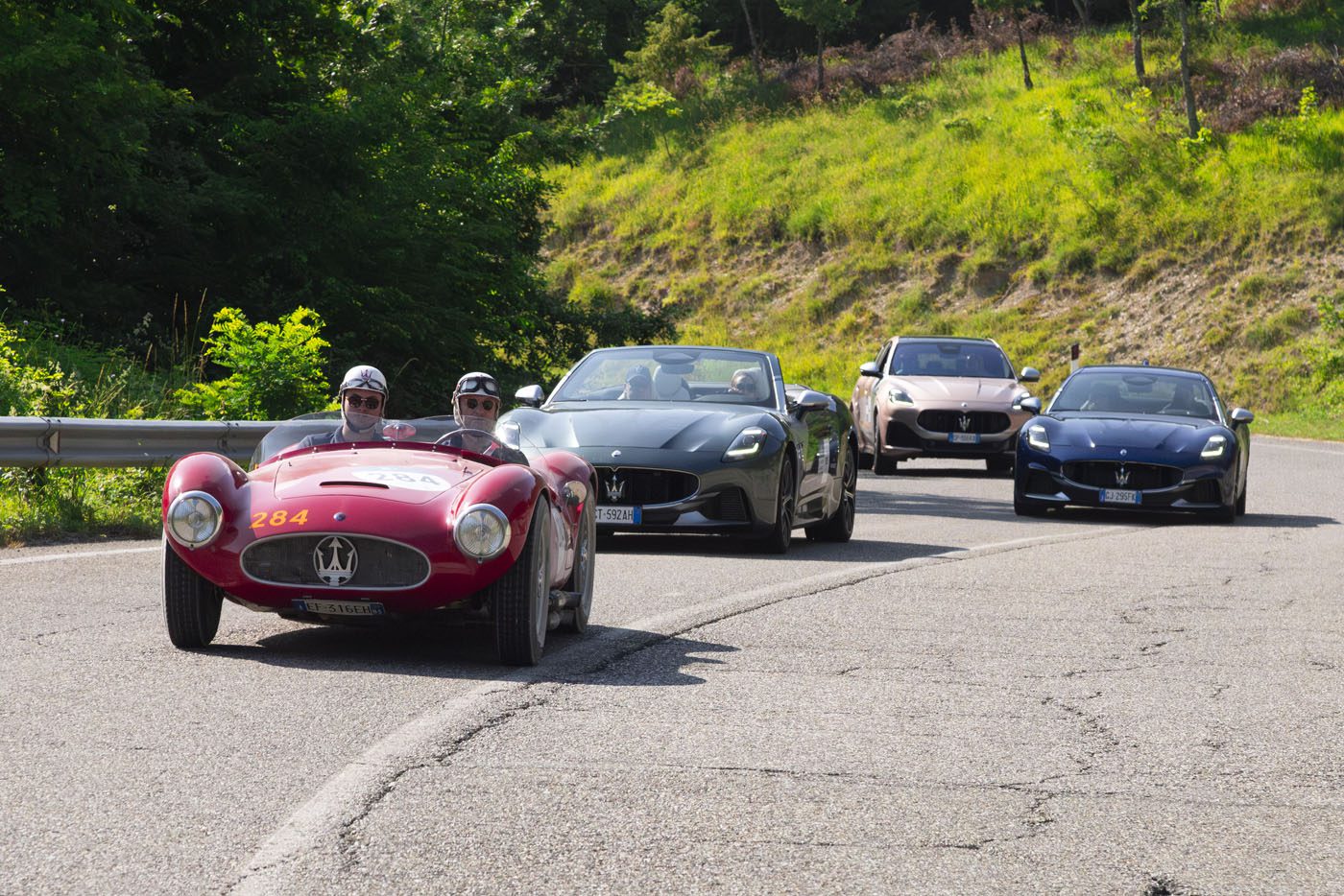 Four Maserati cars, including a vintage red race car reminiscent of the 1000 Miglia and three modern models, drive on a winding road bordered by greenery.