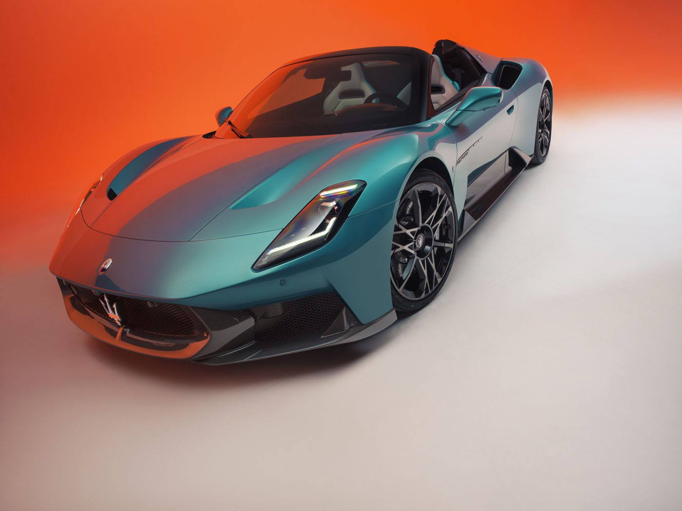 A teal Maserati sports car with the top down is parked on a white surface against a red-orange gradient background.