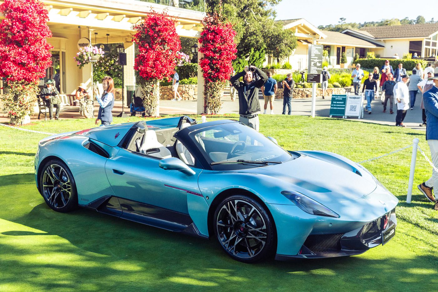 A light blue Maserati GT2 Stradale convertible sports car displayed on a grassy area during Car Week, with people and buildings in the background.