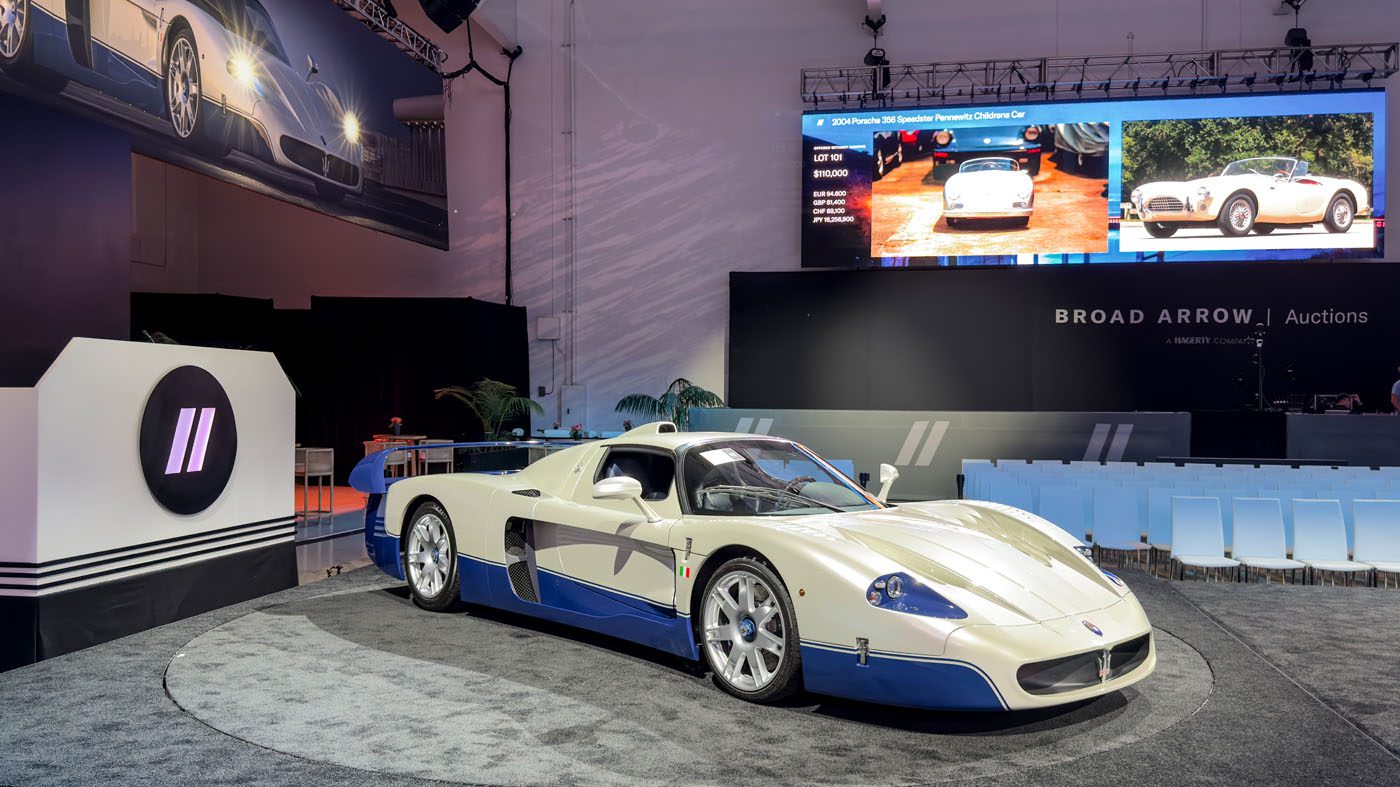 A record-breaking Maserati MC12 auction features a striking white and blue sports car showcased on a round platform, with empty seats and a large screen illuminating the background.