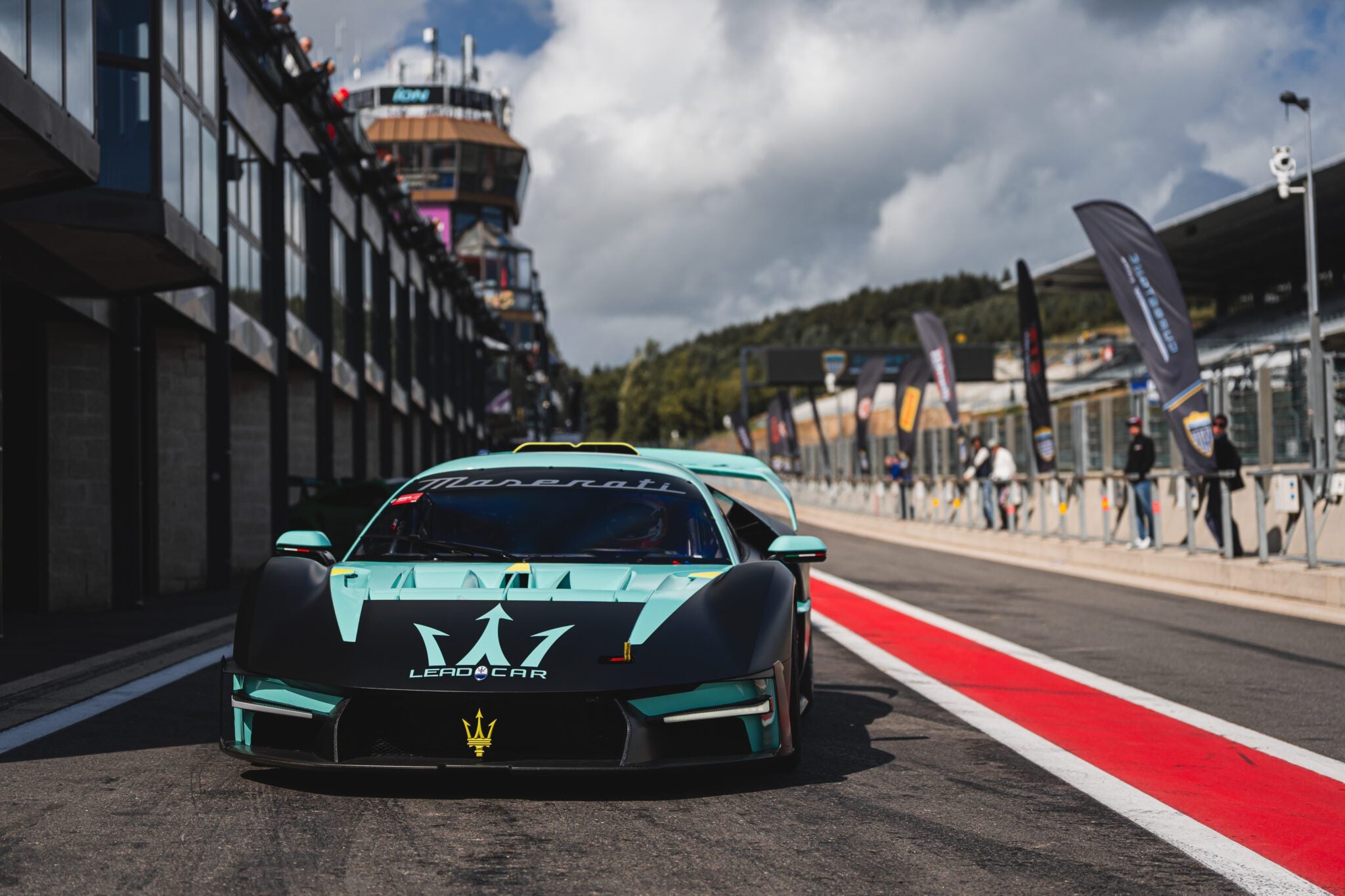 A teal Maserati MCXtrema race car parked in the pit lane at Spa, with another similar car behind it and grandstands rising in the background.