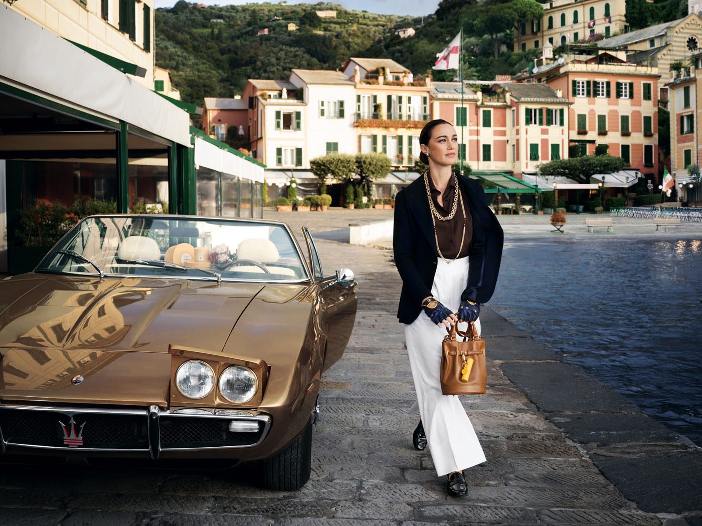 A woman walks beside a vintage brown Maserati parked by a waterfront, with colorful buildings and hills in the background, embodying the elegance of Acqua di Parma’s new collection.