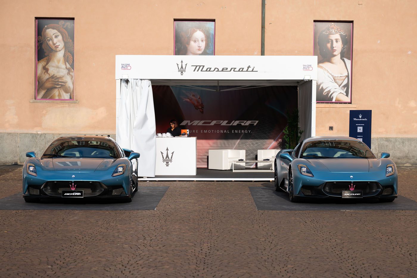 Two blue Maserati MC20 cars are parked in front of a Maserati display booth at the Turin Auto Show, with a person standing inside; two classical paintings adorn the peach-colored wall behind.