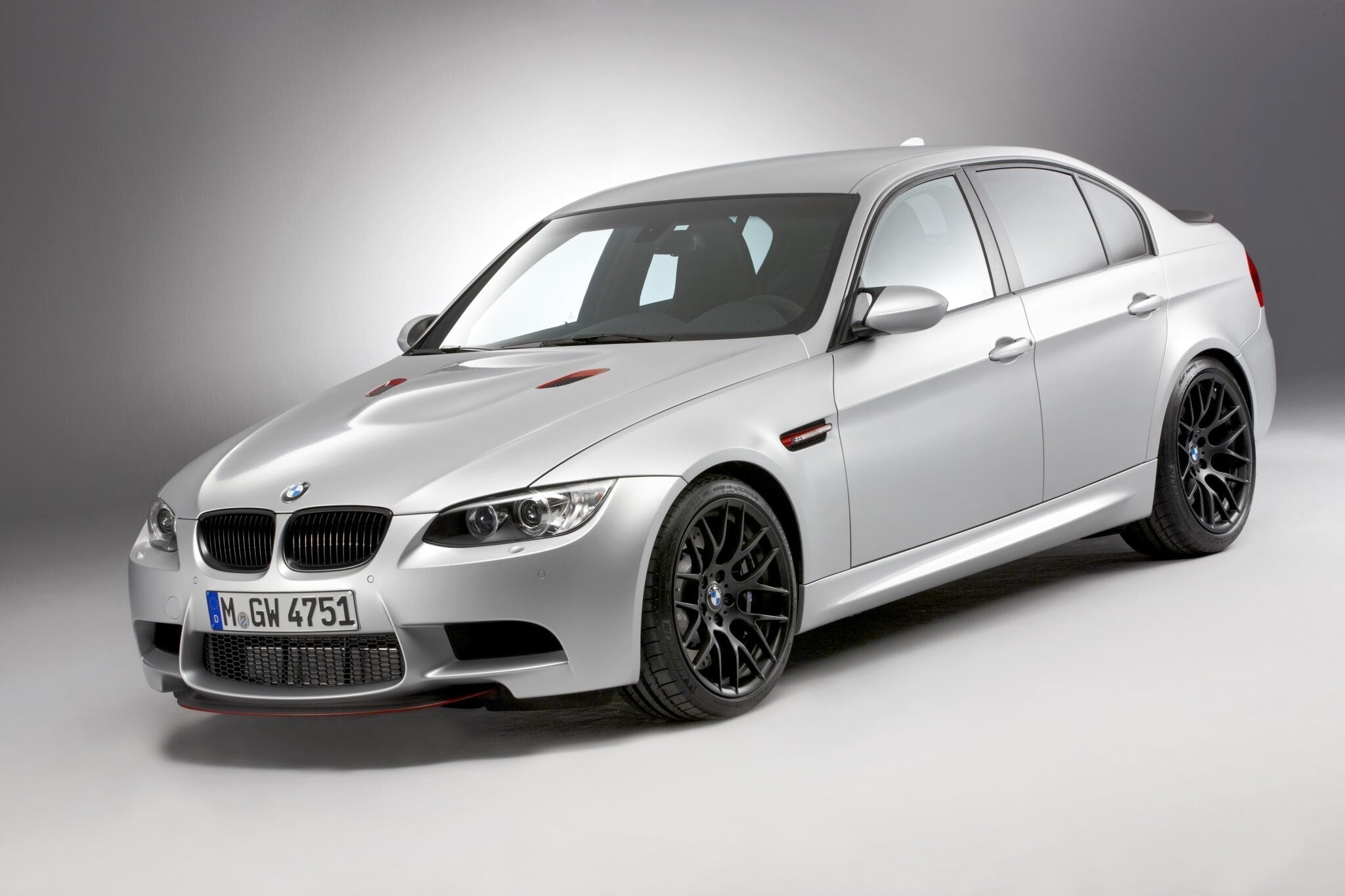 A silver BMW E90 M3 sedan, a true collector car, is parked in a studio with a plain gray background, viewed from a front three-quarter angle.