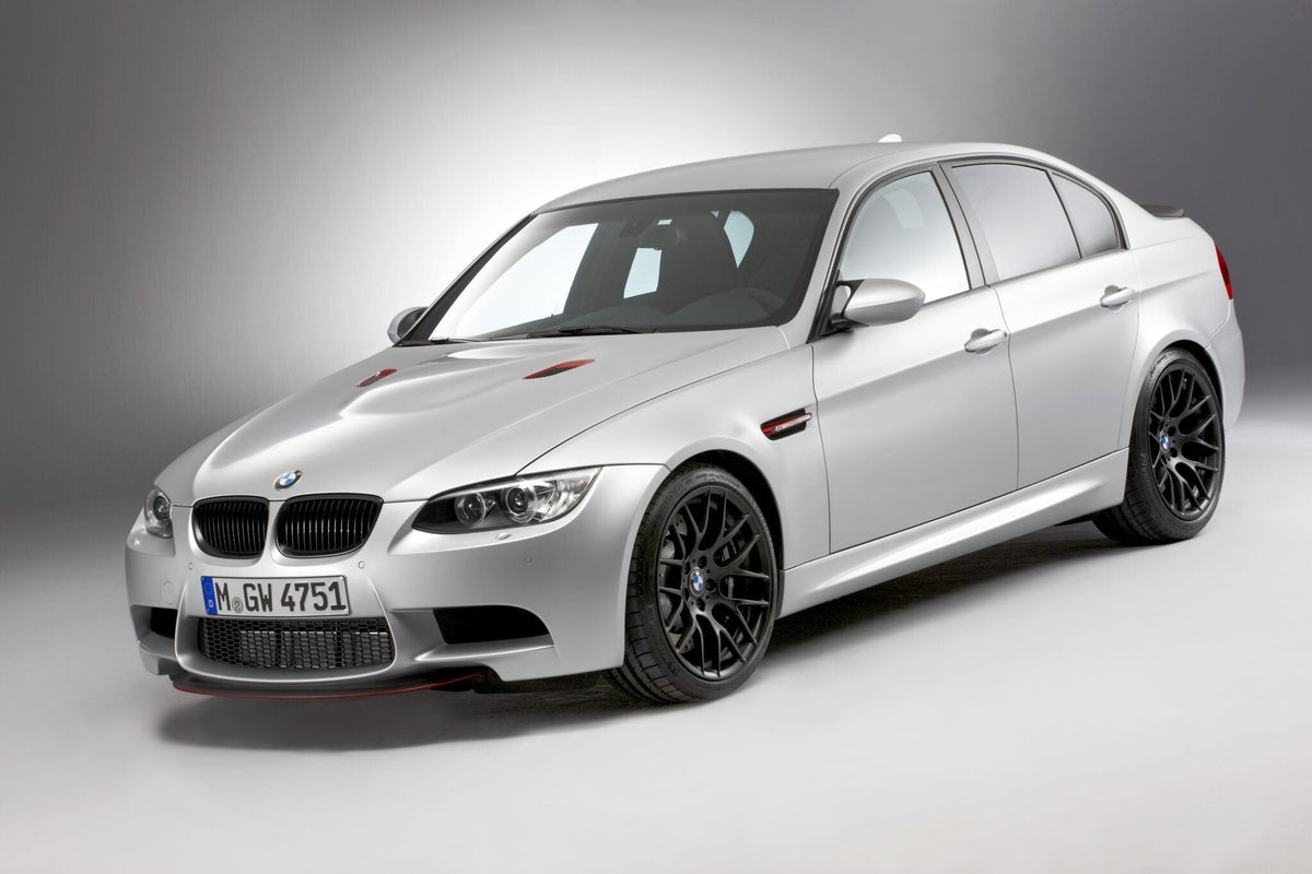 A silver BMW E90 M3 sedan, a true collector car, is parked in a studio with a plain gray background, viewed from a front three-quarter angle.