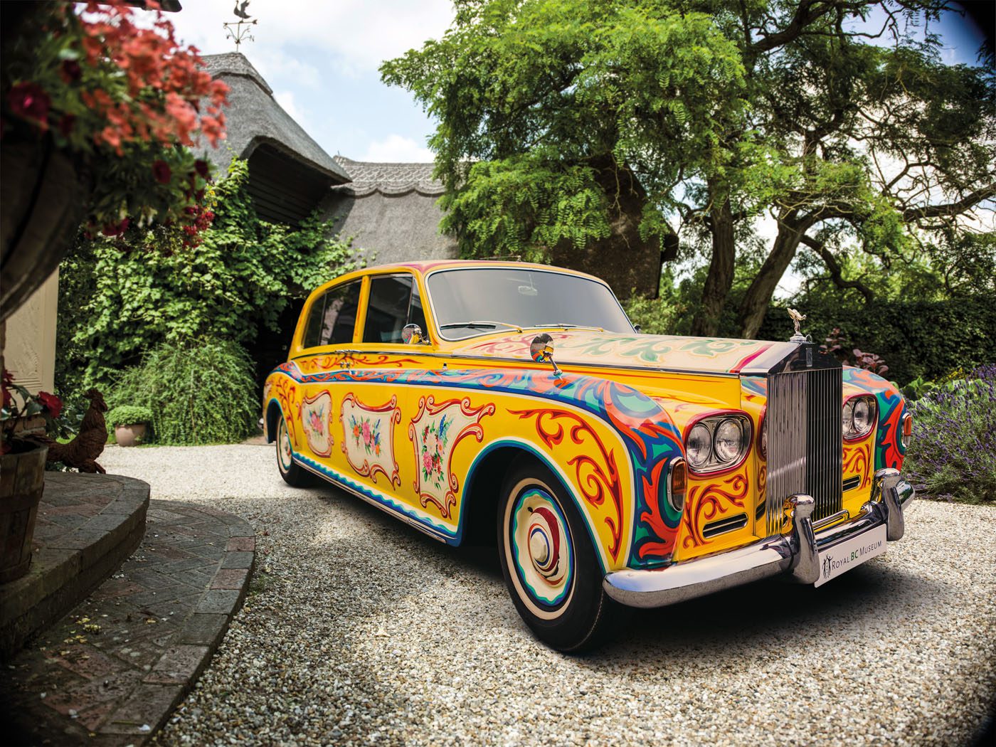 A classic Rolls-Royce Phantom is parked in a gravel driveway, featuring a colorful, psychedelic paint design with intricate patterns and motifs reminiscent of its famous celebrity owners.