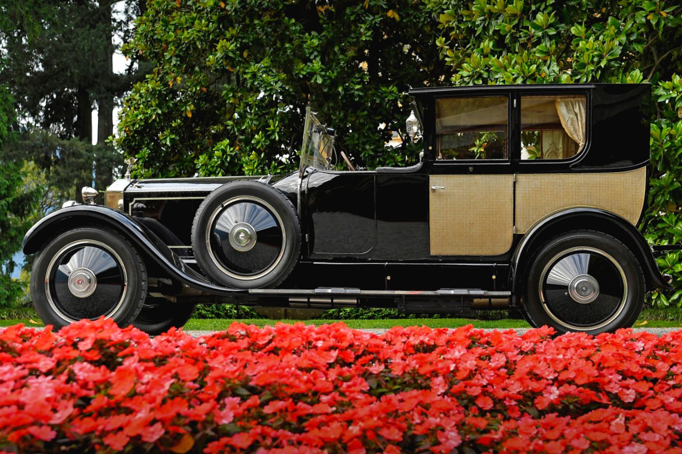 A vintage black and beige Rolls-Royce is parked on a road, with bright red flowers in the foreground and green trees in the background – a scene reminiscent of the elegance found at Goodwood Revival.