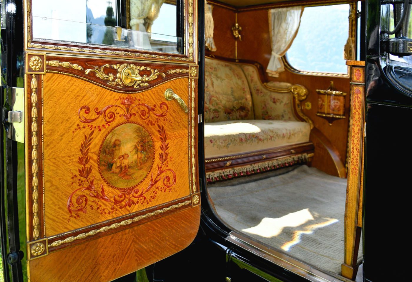 The interior of an ornate vintage carriage with elaborate woodwork, floral upholstery, and decorative painted details on the door evokes the bespoke luxury found in a classic Rolls-Royce Phantom.