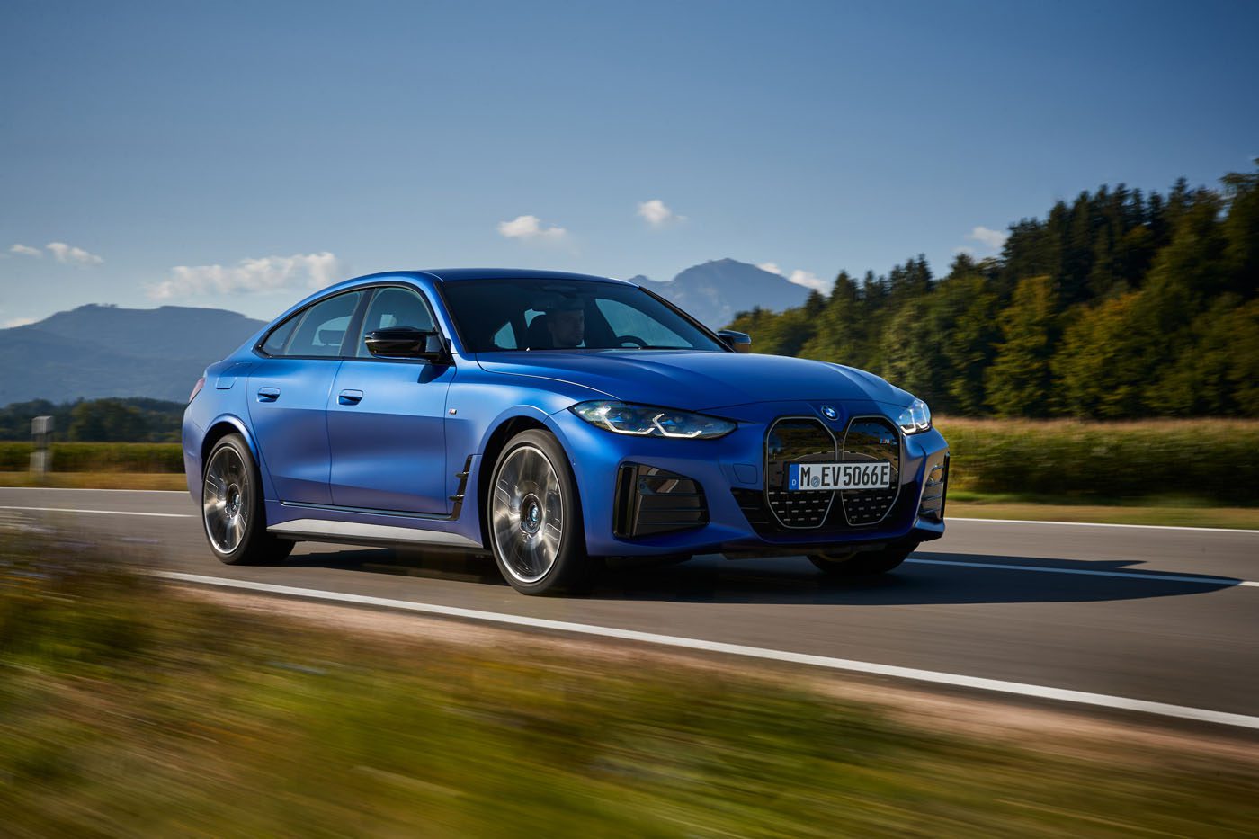A 2024 BMW M blue sedan cruises on a rural road flanked by fields and trees, with majestic mountains in the background under a clear sky, embodying its record sales success.