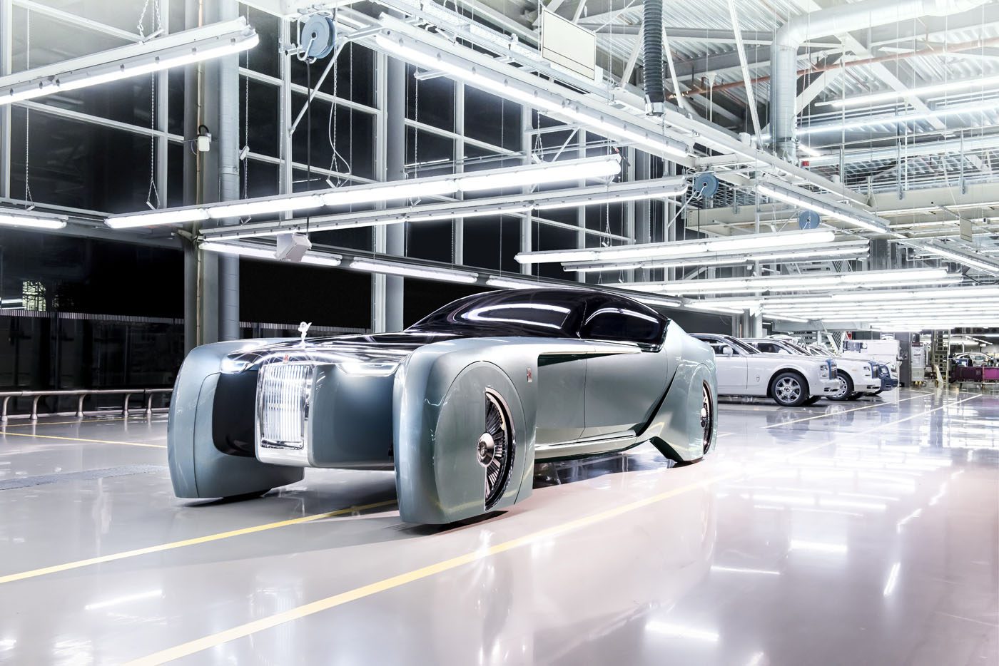 In an industrial showroom, the futuristic concept car stands out with its sleek design and large front grille, reminiscent of the Goodwood Era's elegance. Other cars linger in the backdrop, highlighting its innovation.