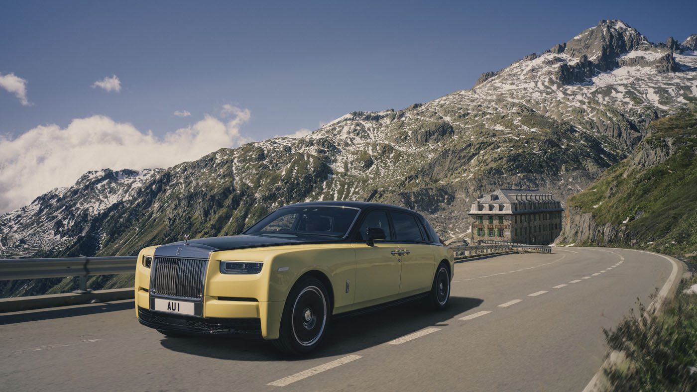 A Rolls-Royce, 007-inspired, glides along a winding mountain road, with rugged, snow-capped peaks in the background under a partly cloudy sky.