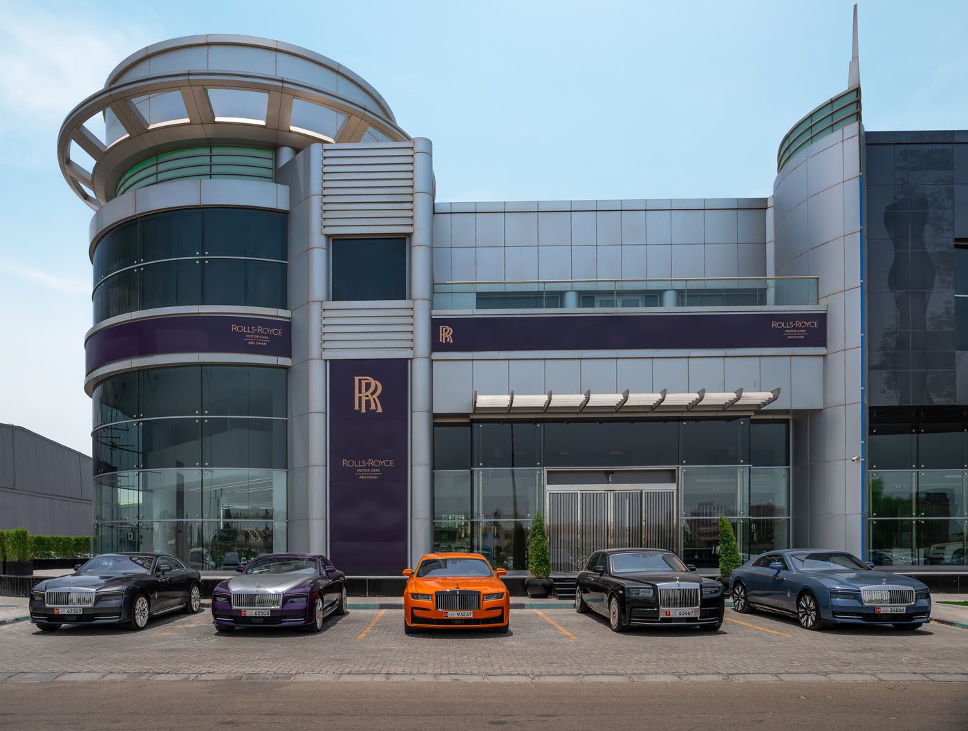 A luxury car dealership in Abu Dhabi showcases five Rolls-Royce cars parked in front of a modern glass and metal showroom.