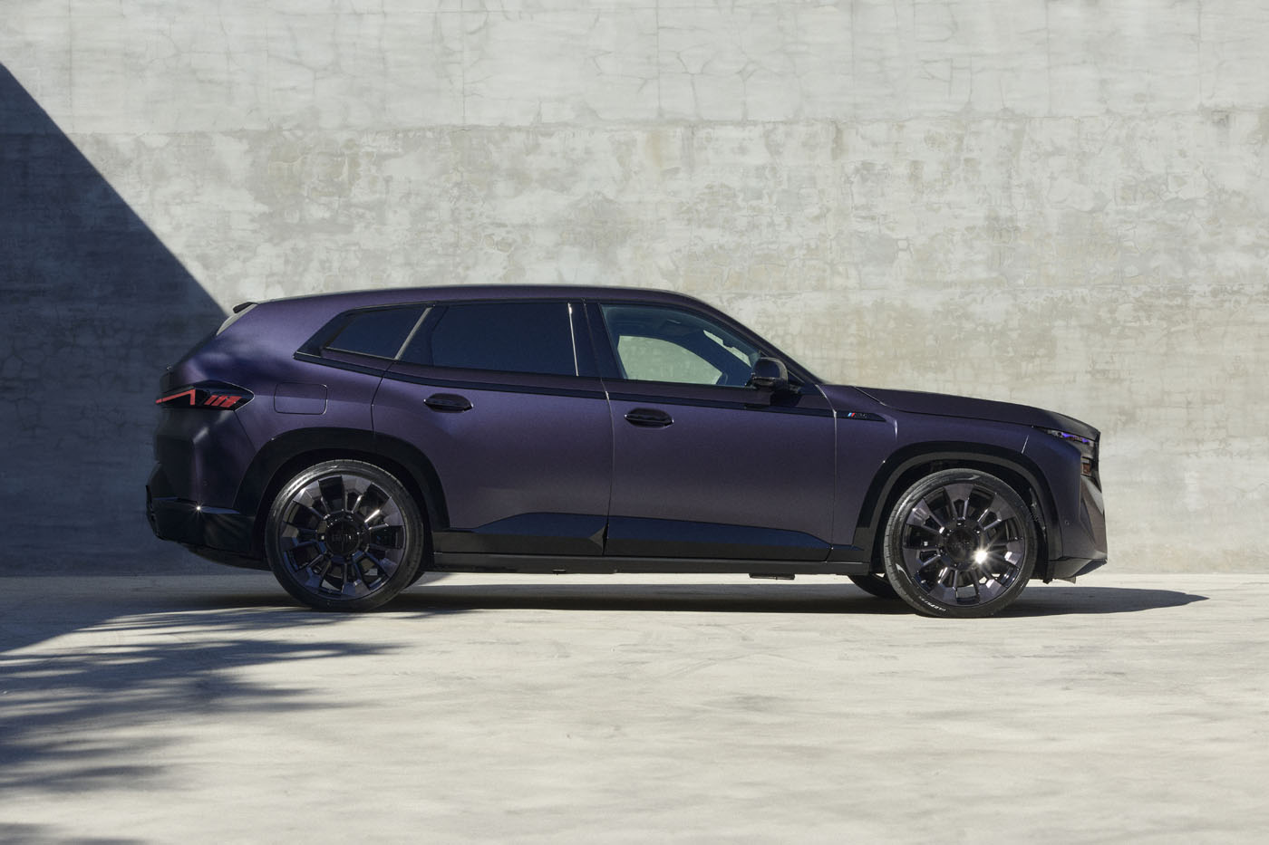 A dark purple 2025 BMW XM Kith Concept SUV is parked on a concrete surface with a textured wall in the background, partially in shadow.
