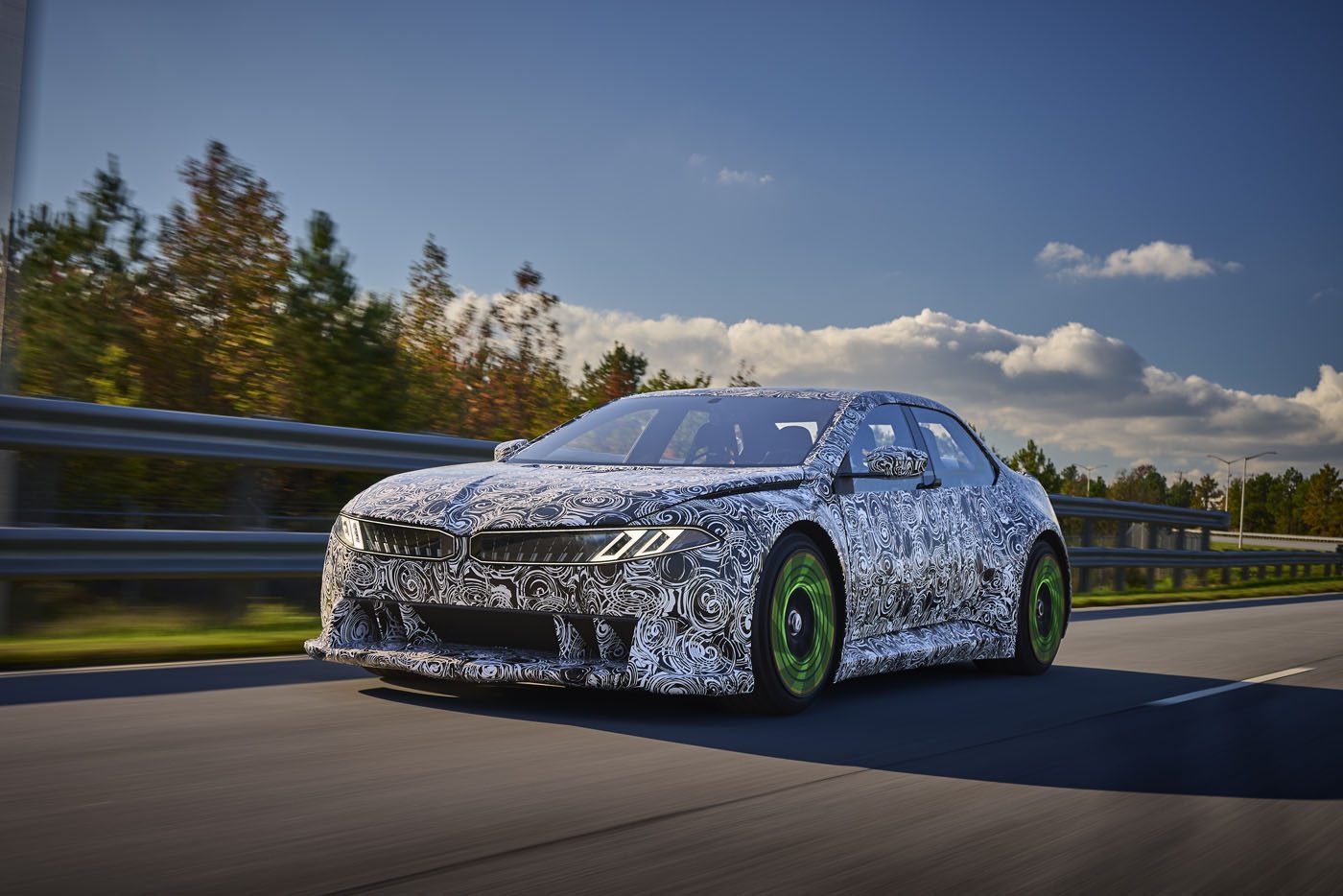 A camouflaged car with green wheels, reminiscent of the BMW Vision design, is driving on a highway surrounded by trees under a partly cloudy sky.
