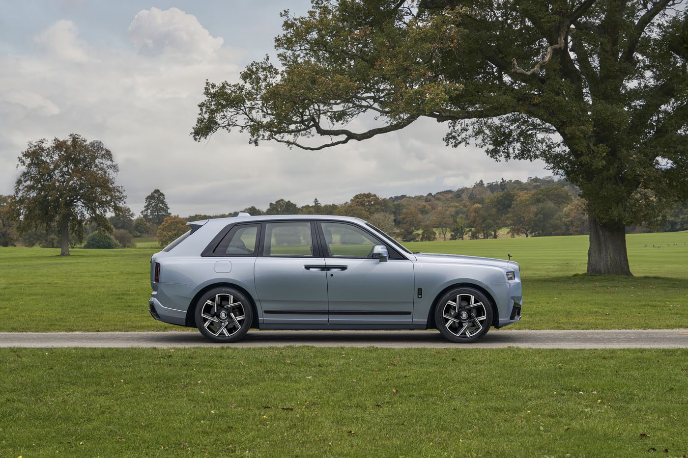 A light blue Rolls-Royce Cullinan is parked on a paved path in a grassy park with large trees and a cloudy sky in the background.