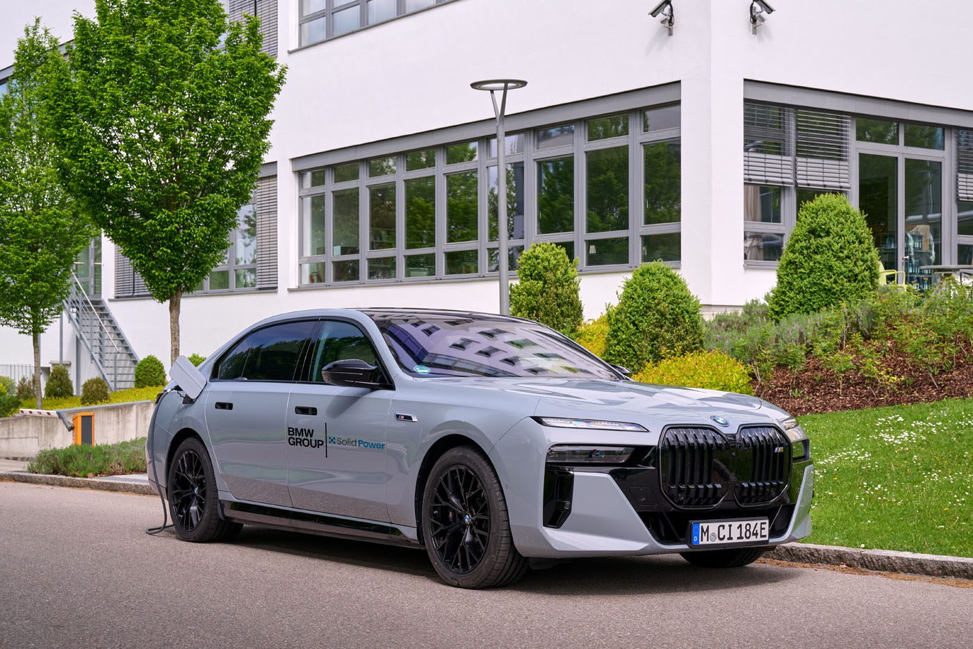 A silver BMW i7 electric sedan is parked by a curb, plugged in for charging, with a modern building and greenery in the background.