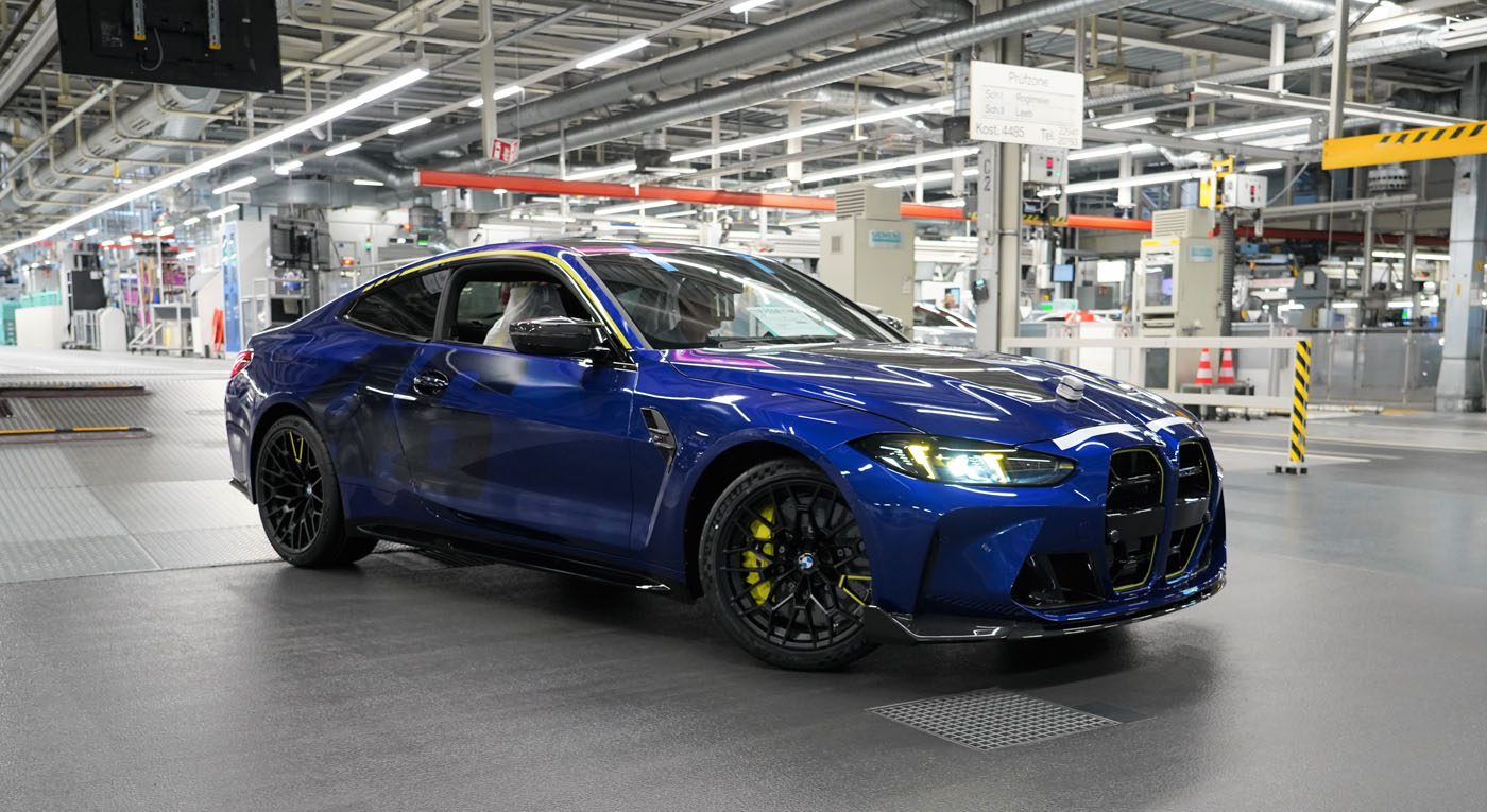 A blue BMW M4 CS coupe parked inside an automotive manufacturing facility, surrounded by industrial equipment and bright overhead lighting, exudes bespoke luxury and engineering excellence.