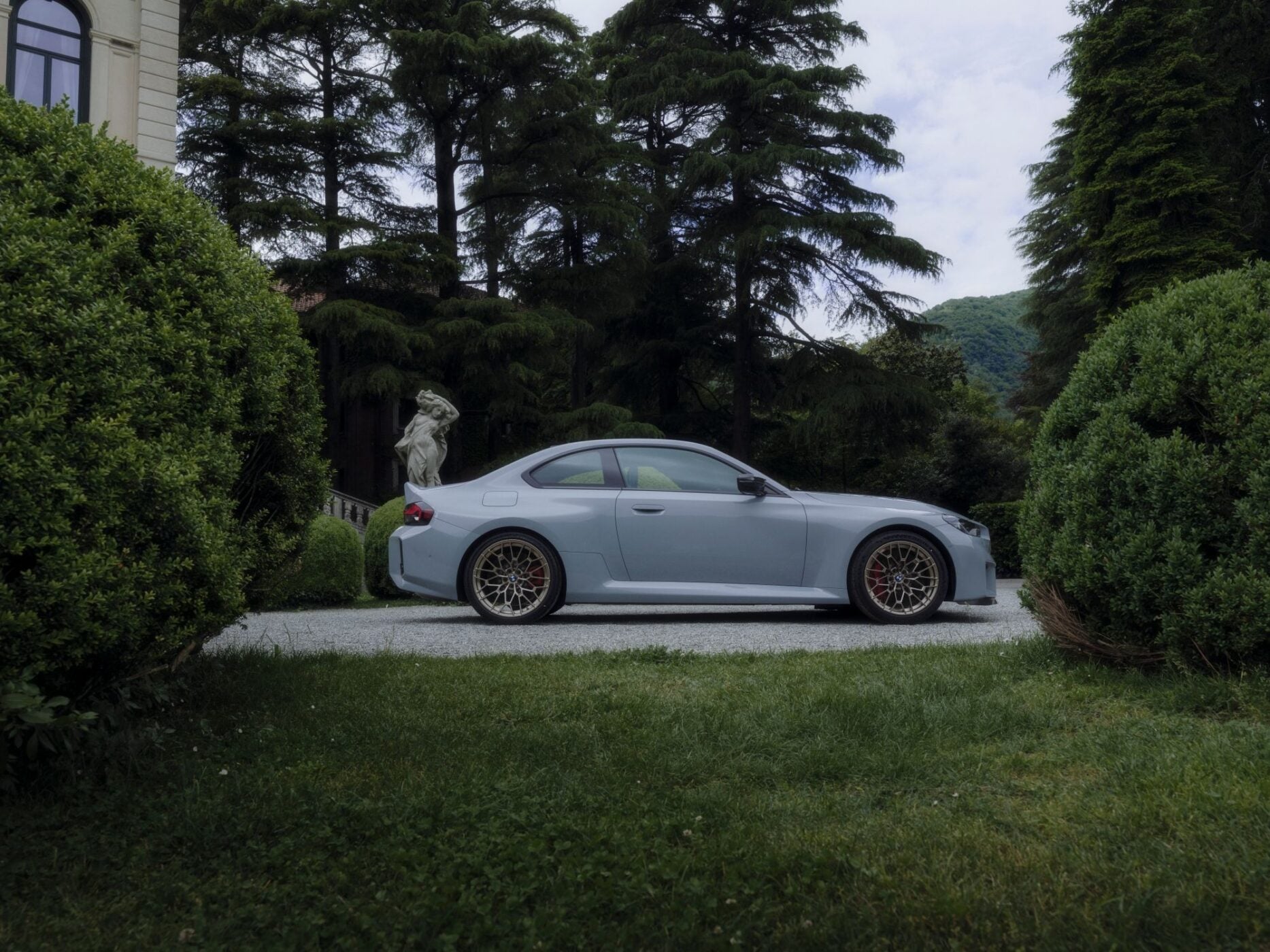 A silver BMW M sports coupe parked on a gravel driveway, surrounded by manicured bushes and tall trees, with a stone statue in the background.