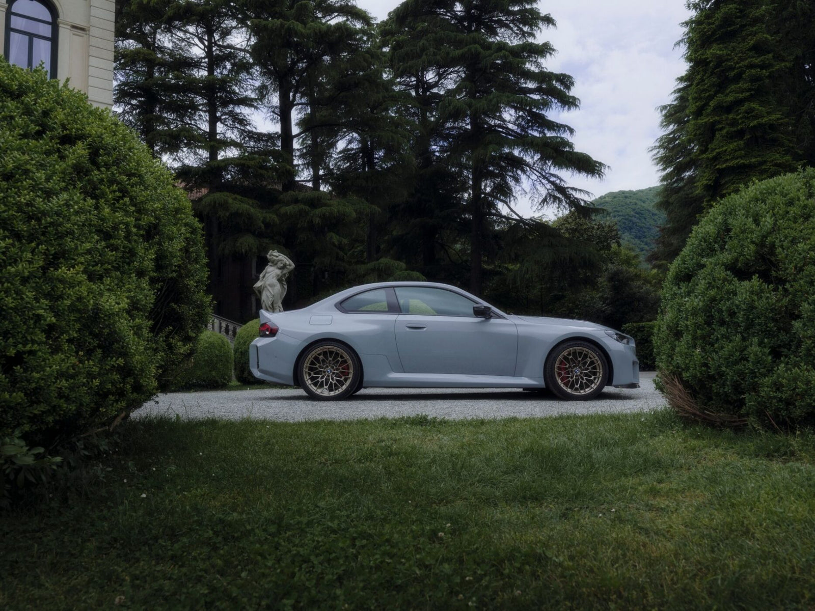 A silver BMW M sports coupe parked on a gravel driveway, surrounded by manicured bushes and tall trees, with a stone statue in the background.