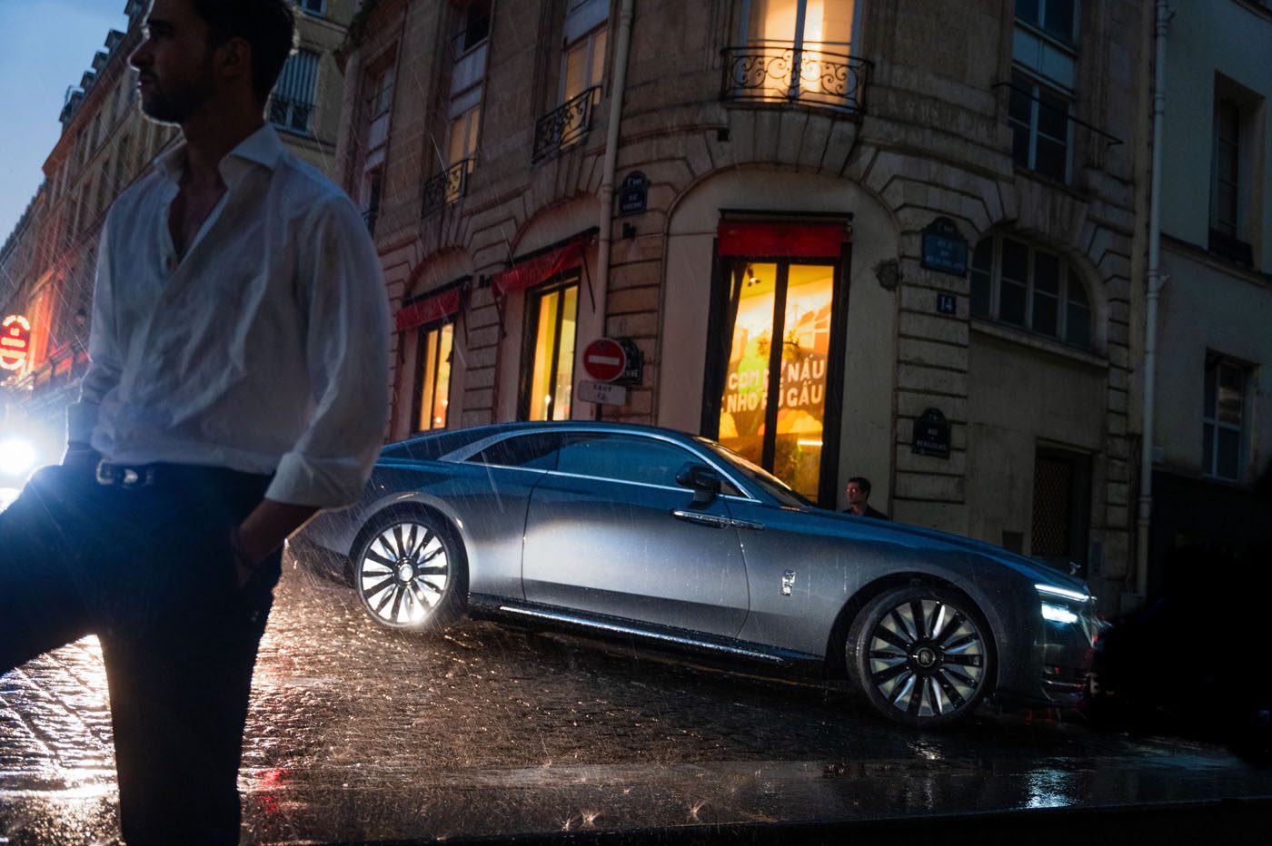 A man in a white shirt stands in the foreground on a wet street, while a gray Cullinan luxury car is parked in front of a building on a rainy evening, evoking the elegance of Paris Fashion Week.