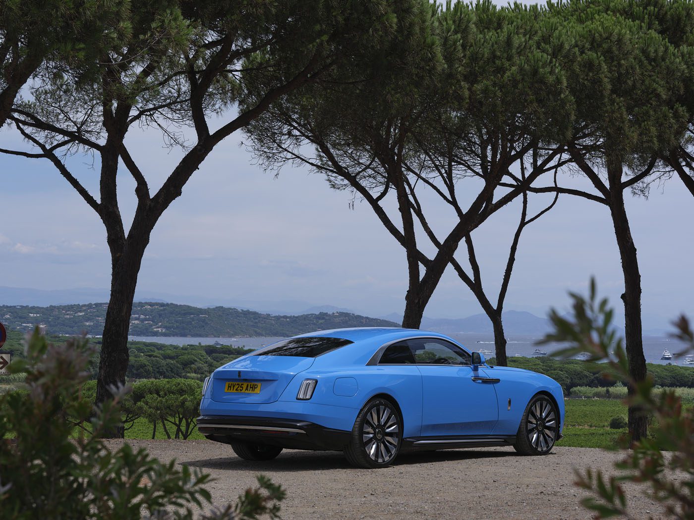 A blue Rolls-Royce luxury coupe with a yellow license plate is parked on a dirt road in Saint-Tropez, surrounded by trees and overlooking a scenic landscape with hills and water.