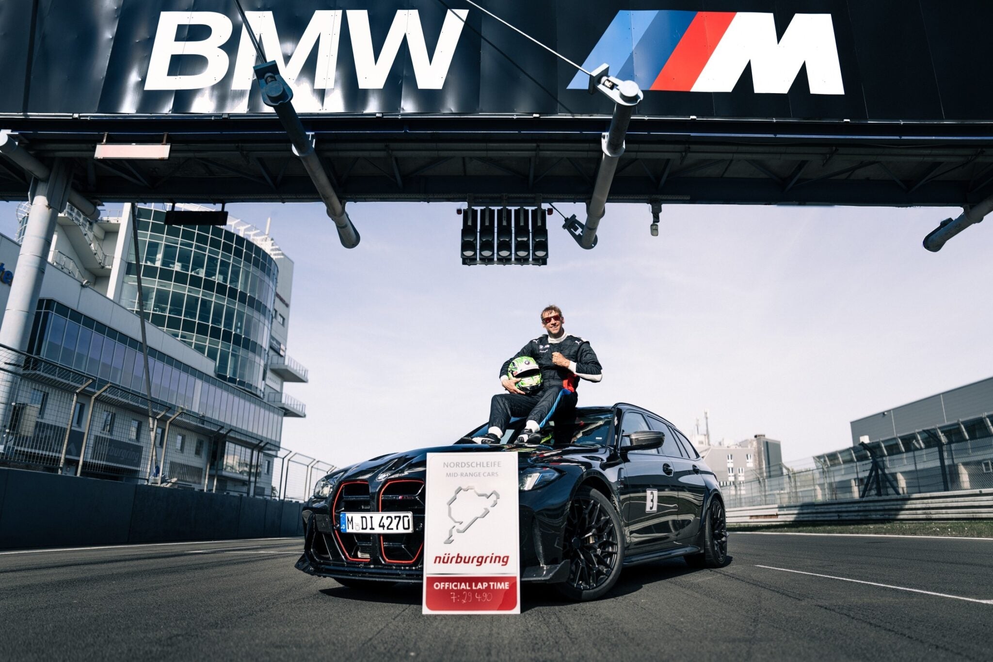 A person in racing gear sits on a black BMW M3 CS Touring under a BMW M sign at a racetrack, with a Nürburgring lap record sign displayed in front, highlighting the fastest wagon’s achievement.
