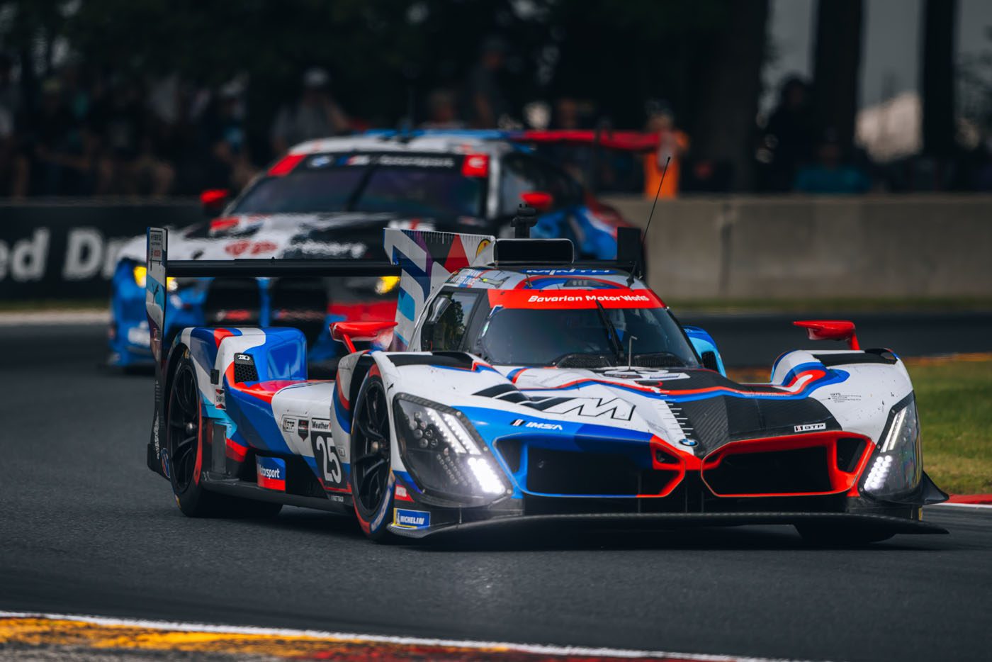 A white, blue, and red race car leads another car on a racetrack during a motorsport event, with spectators and barriers visible in the background.