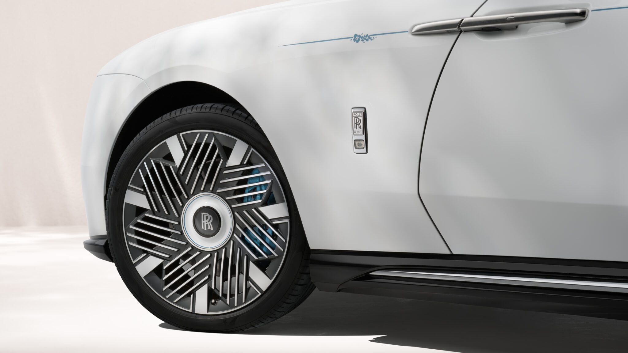 Close-up of a white Rolls-Royce Motor Cars Bespoke Spectre, highlighting the front wheel, unique rim design, and part of the door with a prominent Rolls-Royce badge.