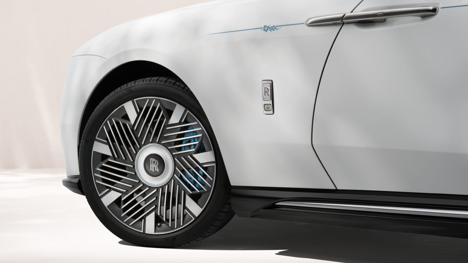 Close-up of a white Rolls-Royce Motor Cars Bespoke Spectre, highlighting the front wheel, unique rim design, and part of the door with a prominent Rolls-Royce badge.