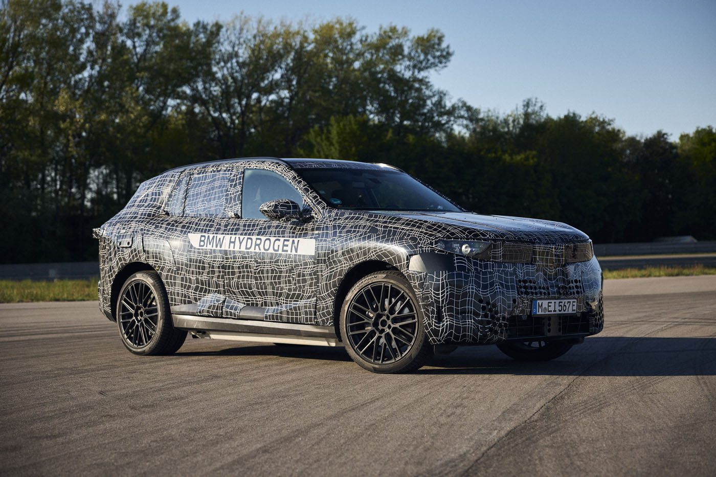 A BMW iX5 SUV test vehicle with a camouflage wrap and “BMW Hydrogen” label is parked on a paved surface in daylight, hinting at hydrogen Series Production, with trees in the background.
