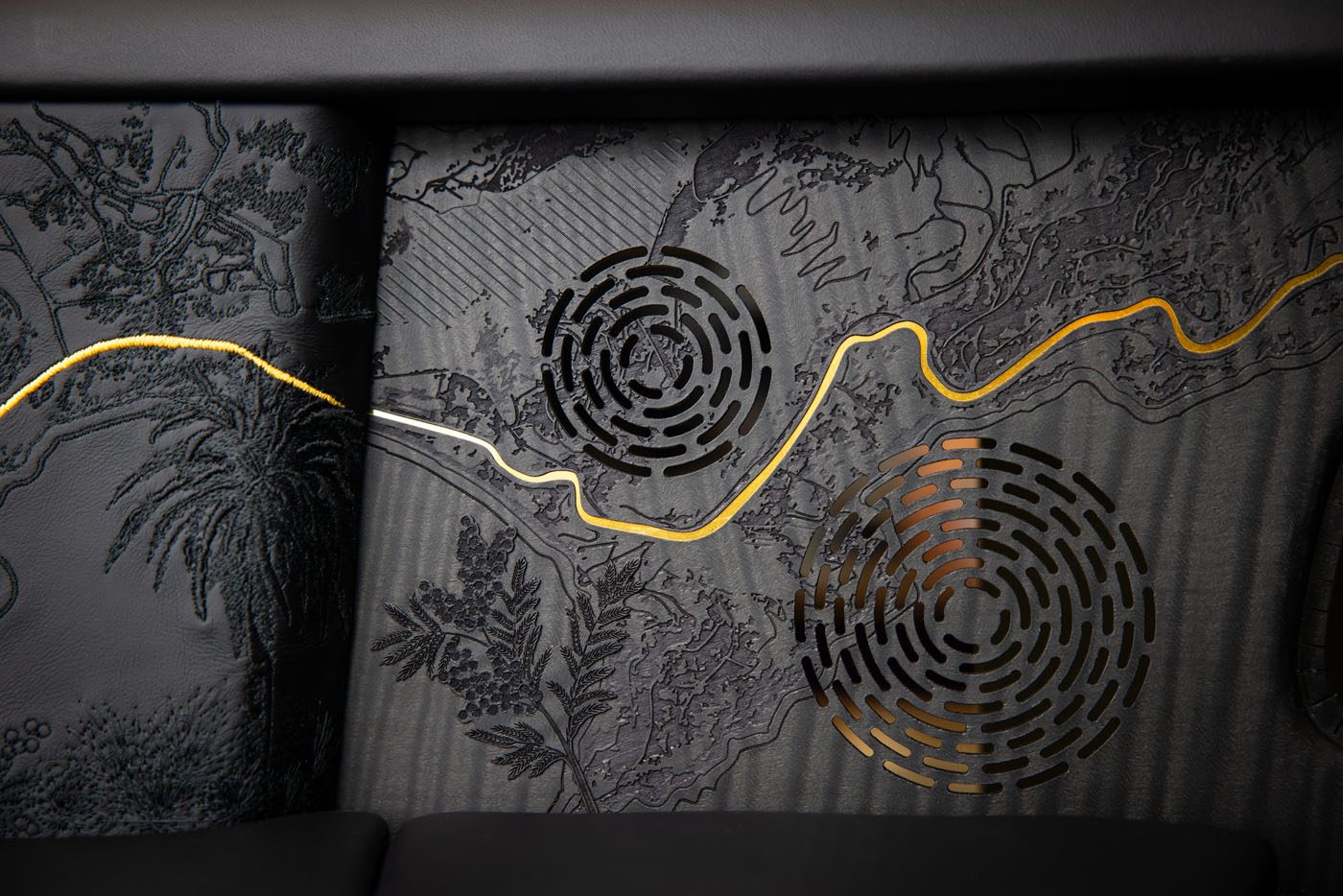 Black textured panel with circular speaker grilles and a wavy gold line, featuring embossed botanical patterns, detailed etching, and intricate woodwork inspired by Rolls-Royce craftsmanship.