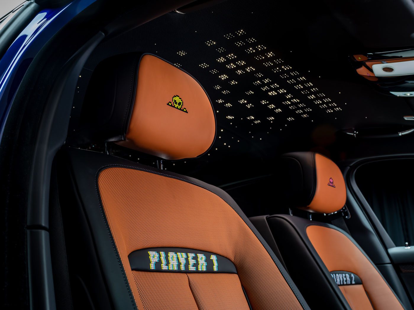 Close-up of two car seats with orange upholstery, embroidered Space Invader icons on the headrests, and "PLAYER 1" and "PLAYER 2" text on the seat backs; starry ceiling above.