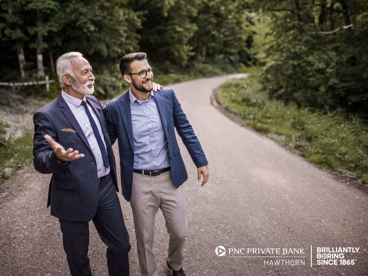 Two men in suits walking and smiling on a tree-lined path, embodying the essence of Generational Wealth. PNC Private Bank and other logos are visible in the corner, emphasizing their commitment to building Lasting Legacies.