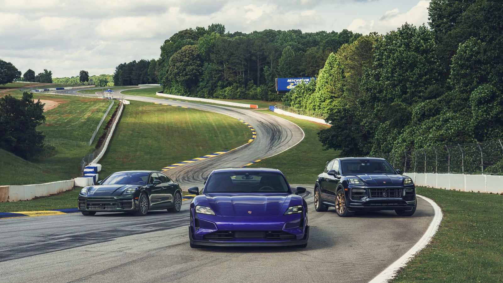 Three Porsche vehicles, including a purple Taycan, are driving on a winding racetrack surrounded by grass and trees.